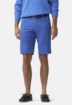 Man draagt een blauwe gebreide trui, blauwe korte broek met patroon van kleine witte ananassen, zwarte riem en zwarte bootschoenen.