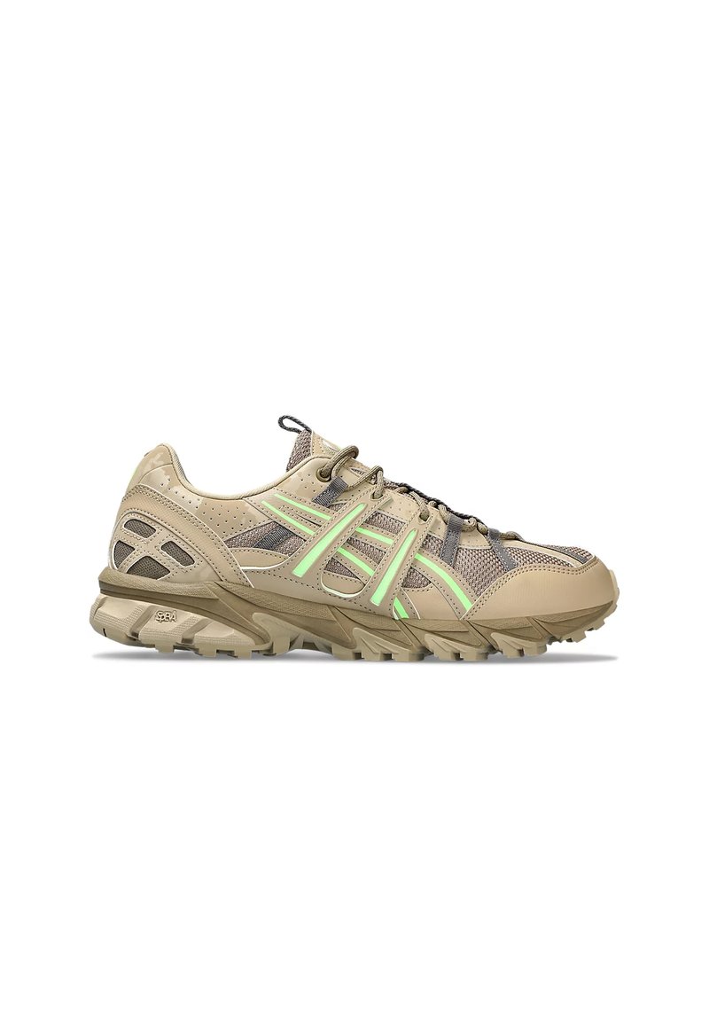 Zapatilla deportiva beige con materiales de malla y sintéticos, con acentos verdes, suela texturizada y áreas reforzadas para mayor durabilidad.