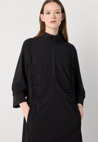 Zwarte athleisure jurk met een hoge kraag, rits aan de voorkant en gerimpelde zijdetails. Bevat zakken en ribgebreide mouwmanchetten. Soepele stoftextuur.