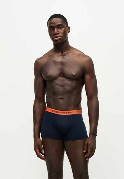 Homme musclé debout portant un boxer bleu marine Emporio Armani avec une ceinture orange sur un fond clair uni.