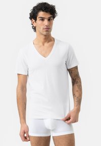 Witte kortarmige V-hals t-shirt gemaakt van soepel stof, gedragen met witte boxershorts. Huid zichtbaar op de armen met tatoeages.