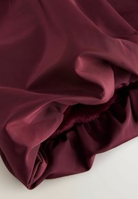 Burgunderfarbene Stofffalten mit einer glatten, leicht glänzenden Oberfläche, die ein flauschiges Innenfutter offenbaren und ein weiches, gerafftes Aussehen erzeugen.