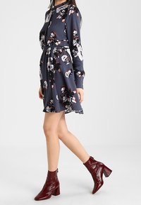 Robe bleu marine à manches longues avec imprimé floral blanc et bordeaux, taille cintrée, associée à des bottines en cuir laqué bordeaux à talons carrés.