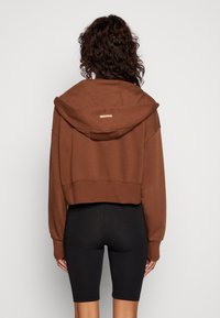 Oříznutá hnědá mikina s kapucí, s detailem zlatého loga na zadní straně. Nošena s černými legínami, které zdůrazňují pohodlný střih.