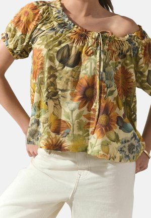 Femme portant un chemisier fleuri épaules nues avec imprimé de tournesols et de feuilles, associé à un pantalon blanc, mains posées sur les hanches.