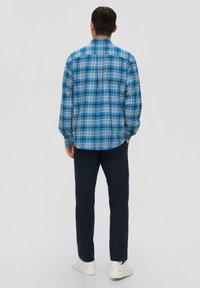 Chemise en coton à carreaux dans des tons de bleu et de noir, à manches longues, col boutonné, portée avec un pantalon bleu marine et des baskets blanches, vue de dos.