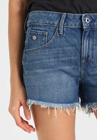 Shorts de mezclilla en azul oscuro con un dobladillo deshilachado. Presenta cinco bolsillos y un cierre de botón metálico. Diseñado con detalles de costura estándar.