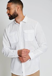 Hombre con barba que lleva una camisa blanca de manga larga abotonada con dos bolsillos en el pecho, de perfil izquierdo, con las manos entrelazadas al frente.