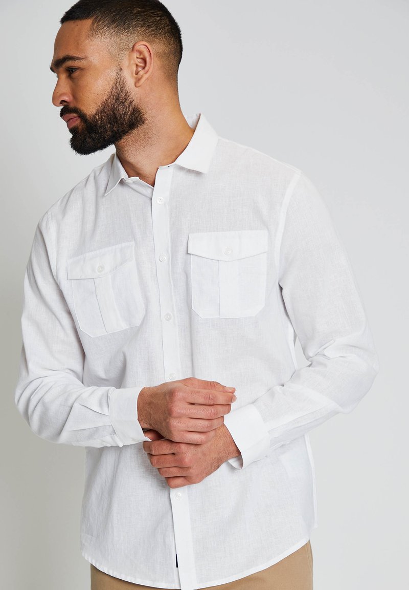 Hombre con barba que lleva una camisa blanca de manga larga abotonada con dos bolsillos en el pecho, de perfil izquierdo, con las manos entrelazadas al frente.