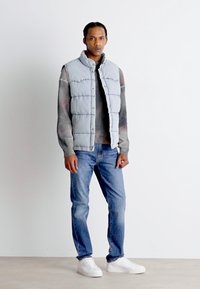 Gilet rembourré gris avec un col montant, pull en tricot gris, jeans bleus et baskets blanches. Texture lisse et coupe décontractée.