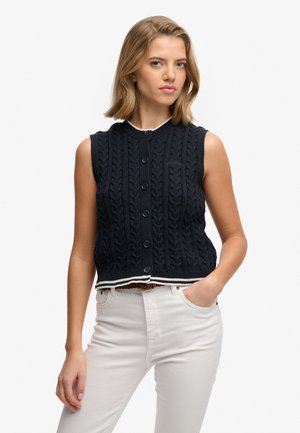 Chaleco sin mangas de punto grueso en azul marino con cierre de botones, con un ribete blanco contrastante en el dobladillo. Textura suave, ajuste informal.