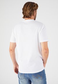 Camisa blanca de punto de manga corta con textura acanalada, cuello redondo y bajo ligeramente más largo en la parte de atrás, combinada con jeans azules con costuras beige.