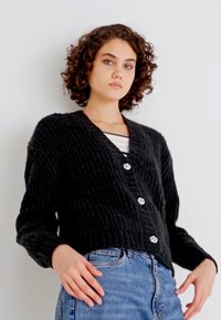 Μαύρο πλεκτό σορτσάκι με βαθύ V-ντεκολτέ και τρία διακοσμητικά κουμπιά, φορεμένο πάνω από ένα λευκό ριγέ μπλουζάκι και συνδυασμένο με τζιν παντελόνι.