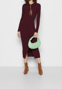 Vestito a coste color bordeaux con bottoni, maniche lunghe, colletto e spacco laterale; abbinato a una borsa verde menta e stivaletti marroni.