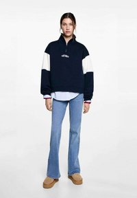 Pull marine avec des manches blanches, col zippé et logo sur le devant, associé à un jean clair évasé et des chaussures à plateforme beige. Texture de tissu lisse.