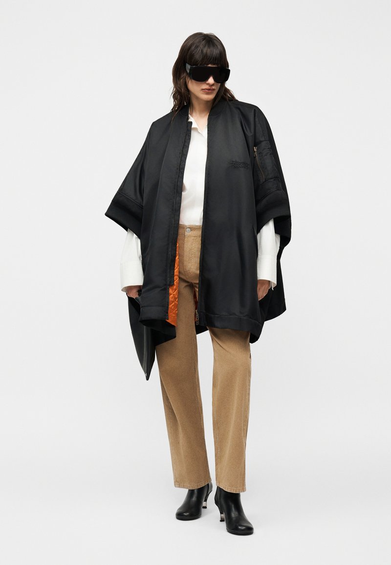 Femme portant une cape noire surdimensionnée avec une doublure orange, une chemise blanche, un pantalon beige, des bottes noires à talons et de grandes lunettes de soleil noires, debout devant un fond blanc.