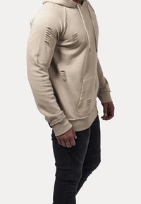 Beige hoodie with a front pocket, featuring distressed details and a drawstring hood, paired with black textured slim-fit pants.