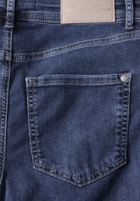 Jean en denim bleu avec une poche arrière unique, présentant une couture renforcée, une texture subtile et une étiquette de marque en cuir au-dessus de la poche.