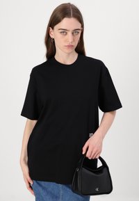 Svart bomull t-skjorte med korte ermer og rund hals, med en liten logo på venstre side. Kombinert med en svart håndveske.