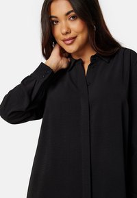 Camicia nera con colletto, maniche lunghe, dettagli in bottone, morbida al tatto e vestibilità comoda, esposta su uno sfondo neutro.