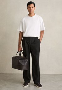 Mann steht und hält eine schwarze Leder-Reisetasche, trägt ein weißes oversized T-Shirt, schwarze weite Hosen und dunkle Sneaker vor einer neutralen Wand.
