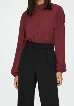 Femme portant un chemisier bordeaux à manches longues avec des boutons et un pantalon noir taille haute à jambes larges sur un fond uni.