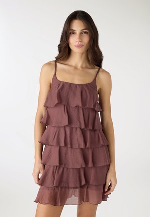 Donna che indossa un vestito marrone senza maniche a balze con sottili spalline, in piedi contro uno sfondo bianco semplice.