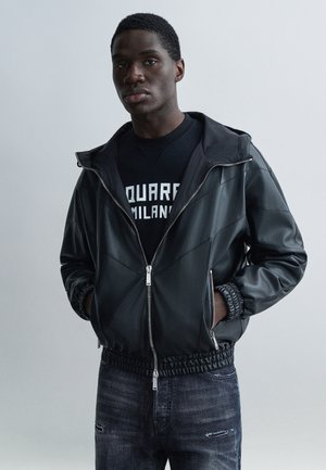 Jonge man met een zwart leren jack over een zwarte trui met witte tekst, staand tegen een effen grijze achtergrond met handen in zijn zakken.