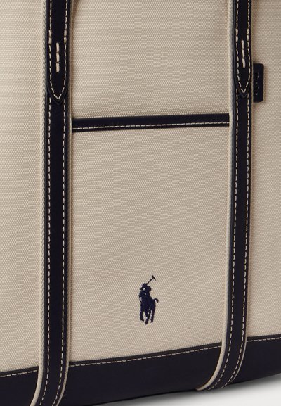 Béžová plátená taška s tmavomodrými koženými detailmi, bielym prešívaním a malým vyšitým logom hráča póla na prednej strane.