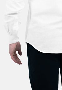 Witte lange-mouwen shirt gemaakt van linnen, met een gebogen zoom en knoopmanchetten, gecombineerd met donkerblauwe broeken, met de hand aan de zijkant geplaatst.