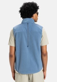 Modrý fleece bez rukávov s vysokým golierom a textúrovanou tkaninou. Zadný dizajn obsahuje horizontálny šev a zavesenú slučku. Viditeľné krátke rukávy.