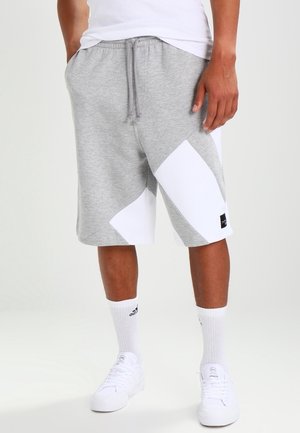 Hombre que lleva pantalones cortos hasta la rodilla en bloques de color gris y blanco, calcetines blancos con logo y zapatillas blancas, de pie con las manos en los bolsillos.