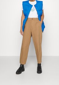Veste matelassée sans manches bleue portée sur un t-shirt blanc avec un texte vert. Pantalon taille haute couleur beige et chaussures noires épaisses. Textures lisses partout.