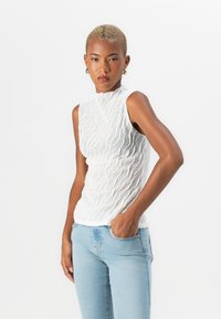 Hvit teksturert ermeløs topp med høy hals, med et bølgete mønster, parret med lyseblå tettsittende jeans.