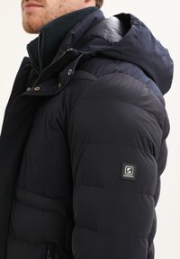 Giacca puffer blu navy con design trapuntato, dotata di cappuccio, chiusura con zip e toppa con logo Gabbiano sulla manica. Tessuto morbido e isolato.