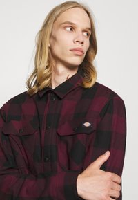 Chemise en flanelle à motifs à carreaux dans les tons de bordeaux et noir, avec deux poches avant, fermeture à boutons et patch de marque sur la poitrine. Texture douce.