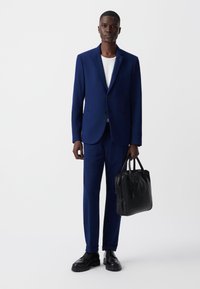 Granatowy garnitur z dopasowaną marynarką i pasującymi spodniami. Model trzyma czarną skórzaną torbę. Czarne buty dopełniają wygląd.