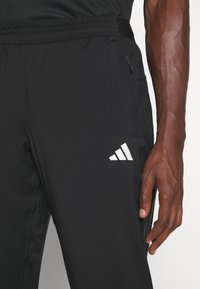 Svarta träningsbyxor tillverkade av lättviktig textil, med elastisk midja, sidofickor och en vit Adidas-logga på låret.