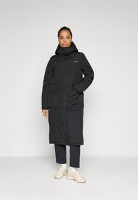 Woman standing in black long padded coat with hood, black pants, and beige sneakers against a plain light background.