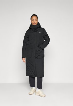 Woman standing in black long padded coat with hood, black pants, and beige sneakers against a plain light background.