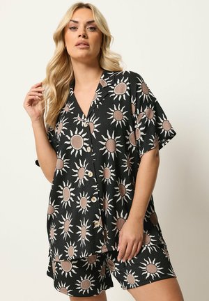 Kvinde iført sort knap-op skjorte og shorts med brune solmønstre, poserende mod en ensfarvet lys baggrund.
