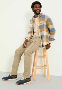 Karierte Flanellshirt in Blau und Gelb, beige Cargo-Hosen und blaue Sneakers mit braunen Akzenten, sitzend auf einem Holzschemel.