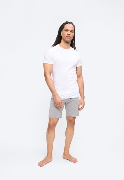 Άνδρας με μακριά μαλλιά που φοράει λευκό εφαρμοστό μπλουζάκι και γκρι σορτς, όρθιος ξυπόλητος σε απλό φωτεινό φόντο.