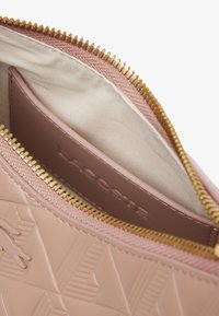 Intérieur de sac à main rose texturé avec une fermeture éclair dorée, une doublure beige et un porte-cartes marron avec "LACOSTE" embossé.