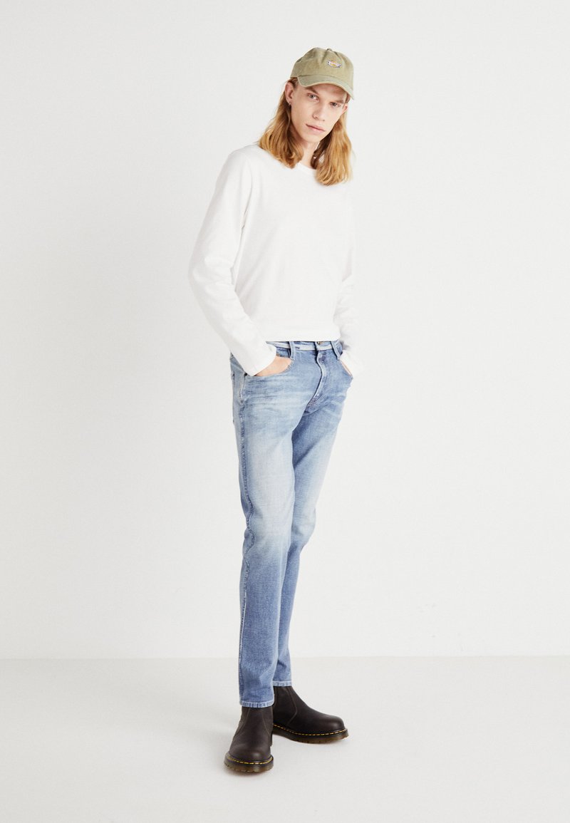 Weißes Langarmshirt, hellblaue Jeans mit ausgeblichenen Details, dunkelbraune Stiefeletten mit gelben Nähten und eine hellgrüne Mütze.