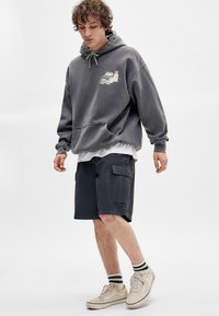 Un sweat à capuche gris avec une poche avant et un design graphique, associé à un short cargo noir. Des baskets beiges avec des chaussettes à rayures vertes complètent l'ensemble.