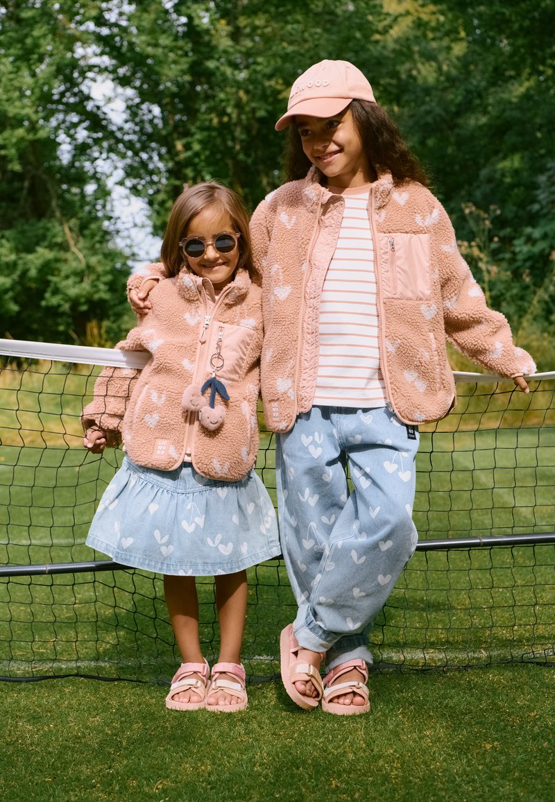 Δύο παιδιά στέκονται δίπλα σε ένα δίχτυ τένις. Το μεγαλύτερο φοράει ροζ fleece μπουφάν με σχέδια καρδιών και ριγέ πουκάμισο· το μικρότερο φούστα από τζιν και ροζ σανδάλια.