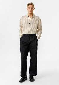 Crème- en grijs gestreepte blouse met lange mouwen en een borstzak, gecombineerd met hoge zwarte broeken en zwarte veterschoenen.