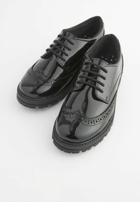 Chaussures en cuir verni noir avec un design brogue, présentant des perforations décoratives, des lacets ronds et une semelle en caoutchouc texturée.
