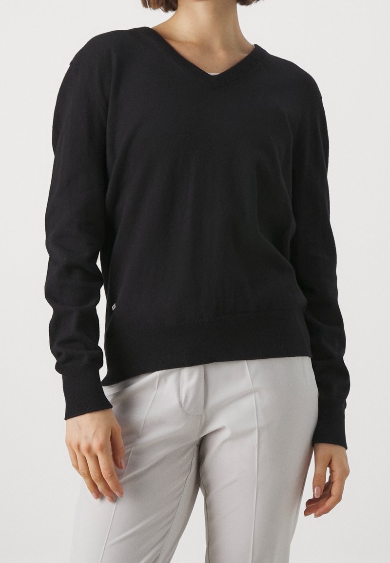 Femme portant un pull noir à col en V à manches longues et un pantalon plissé gris clair, debout avec les mains détendues sur les côtés.